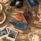 Three personalized leather luggage tags in blue, brown, and tan are arranged on a rustic wooden table with travel-themed props including a camera, compass, passport, coins, rope, and polaroid photos.