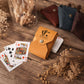 A tan leather playing card holder with a silver snap button and a custom 'HENRY' logo printed on the front, shown on a rustic wooden table next to playing cards and surrounded by dried flowers. Other card holders in brown and blue are visible in the background.