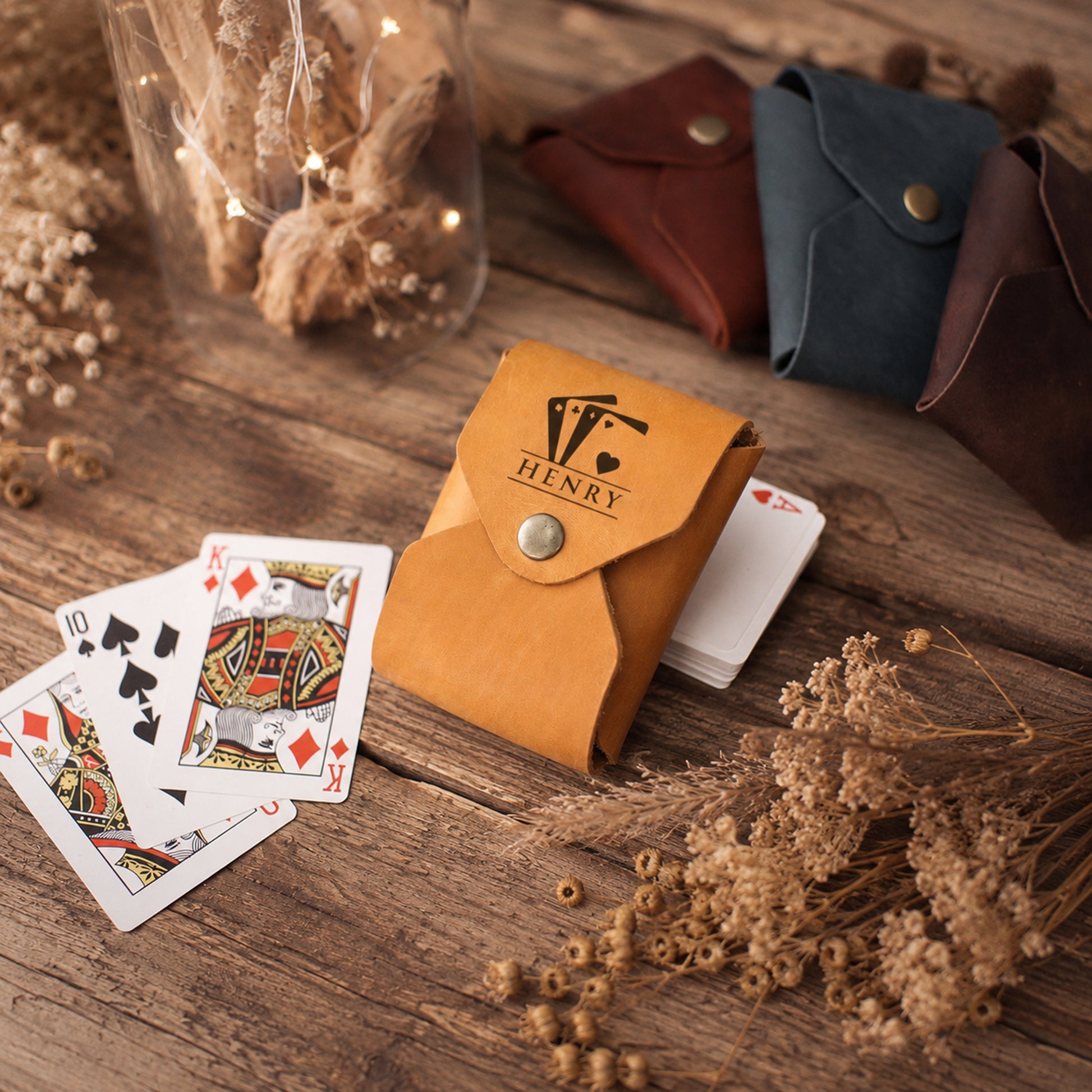 A tan leather playing card holder with a silver snap button and a custom 'HENRY' logo printed on the front, shown on a rustic wooden table next to playing cards and surrounded by dried flowers. Other card holders in brown and blue are visible in the background.