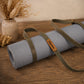 A rolled light gray yoga mat with olive green straps and a brown leather brand tag reading 'Chicotte', displayed on a wooden tabletop with dried flowers in the background.