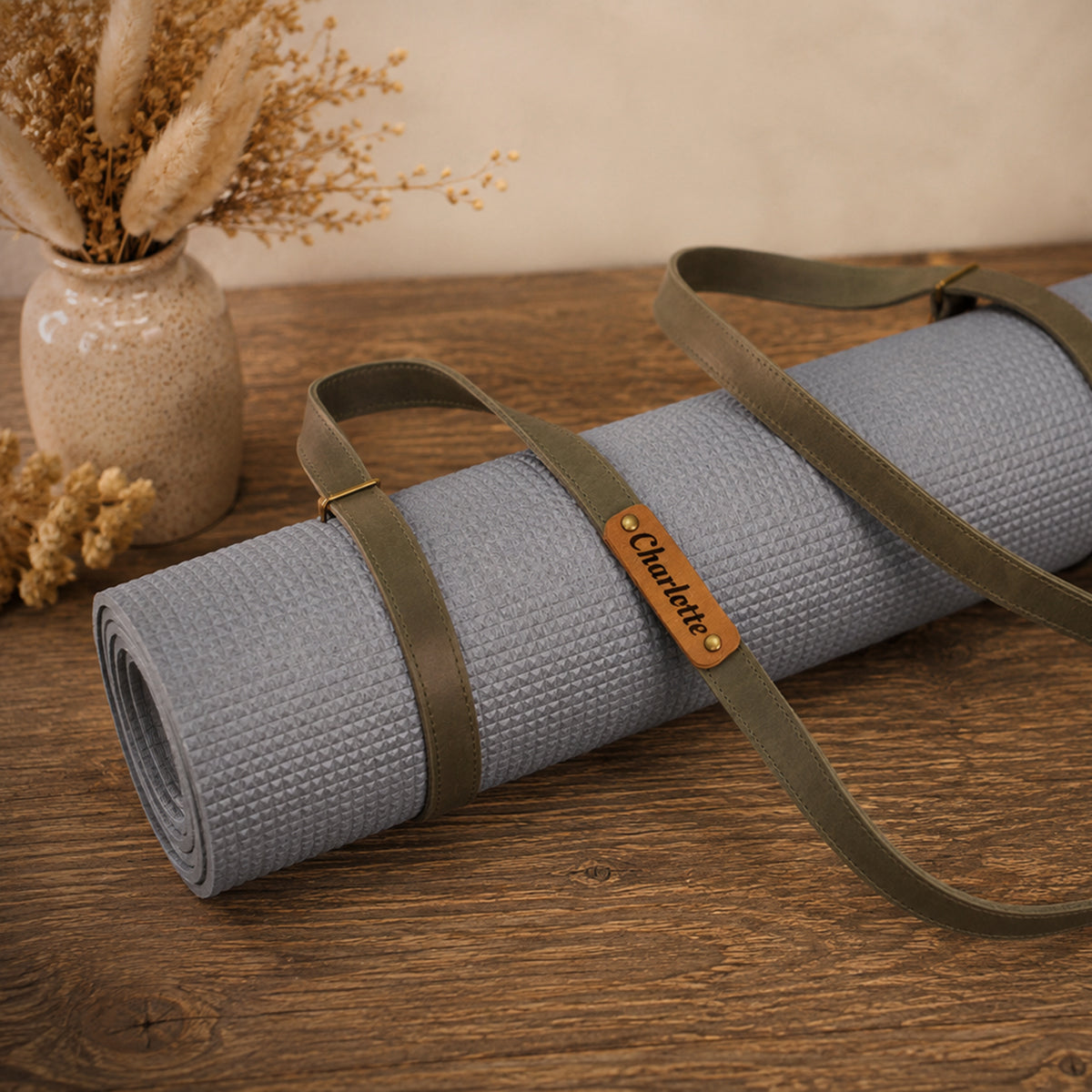 A rolled light gray yoga mat with olive green straps and a brown leather brand tag reading 'Chicotte', displayed on a wooden tabletop with dried flowers in the background.