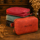 A stack of three zippered pouches or small cases sits on a wooden table, with a coral-red front pouch labeled OLIVER in raised lettering. The top pouch is a muted red and the middle pouch is pale blue-green; a black pouch is partially visible beneath.