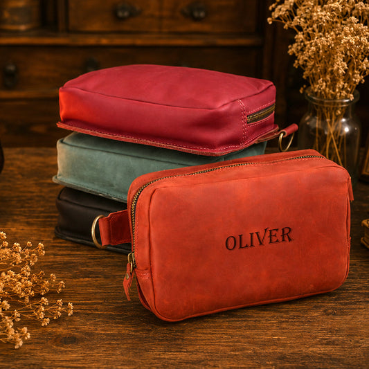 A stack of three zippered pouches or small cases sits on a wooden table, with a coral-red front pouch labeled OLIVER in raised lettering. The top pouch is a muted red and the middle pouch is pale blue-green; a black pouch is partially visible beneath.