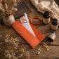 An orange suede pill organizer case with a clear plastic compartment and white cap, styled on a rustic wooden table with dried flowers, a book, a cream knit blanket, and three supplement bottles.