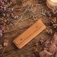A tan leather wallet or card holder with snap closures, styled on a rustic wooden surface with dried flowers, jewelry, and a candle.