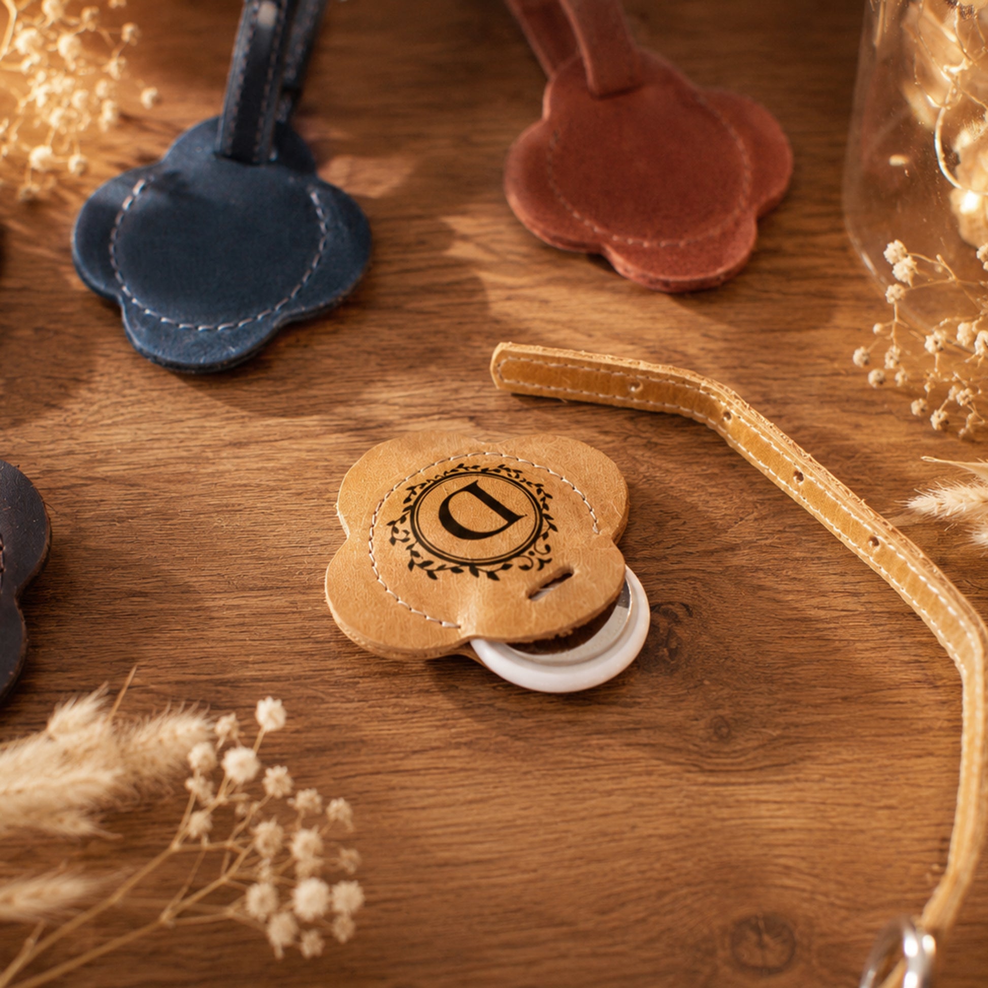 A close-up of a tan leather pet collar with a round flower-shaped metal tag engraved with a monogram, resting on a wooden surface with dried flowers and other collars in the background.