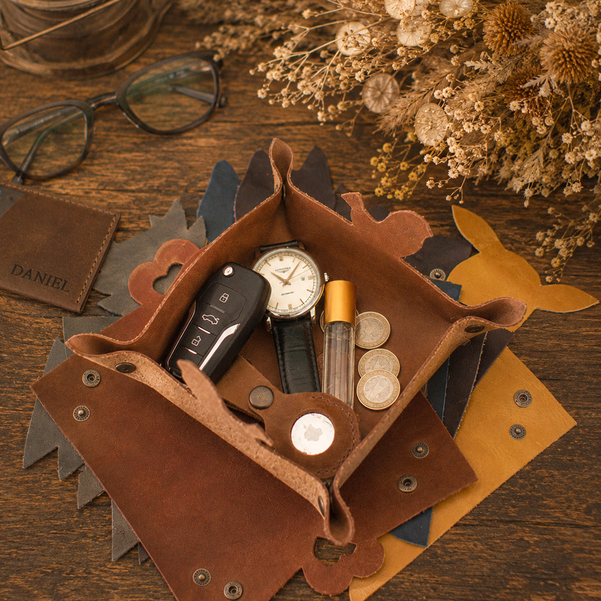 A brown leather organizer tray with scalloped edges and metal snap details, styled on a wooden tabletop with dried flowers, eyeglasses, a wallet, a watch, coins, and a small bottle. The tray is open and contains these items as props.