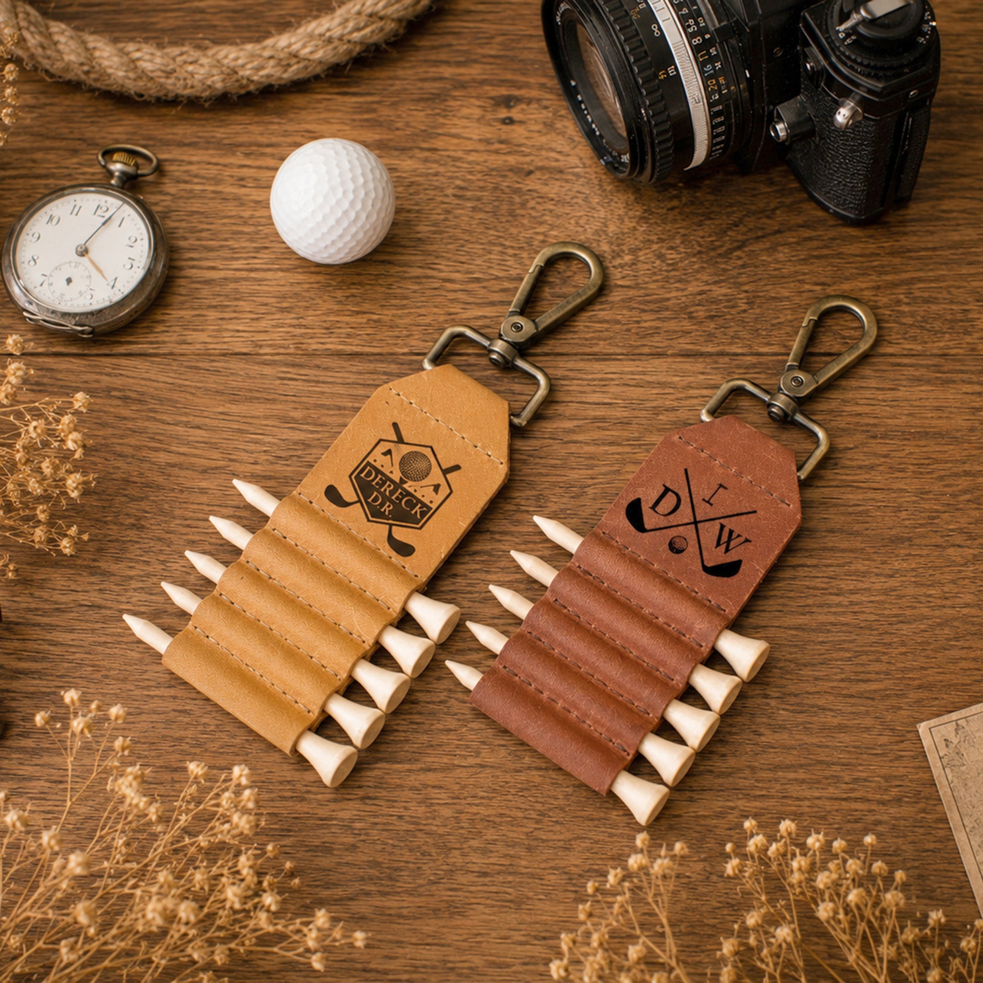 Two leather golf keychains styled on a wooden tabletop with vintage props including a pocket watch, camera, golf ball, and dried flowers. One keychain is tan and the other is brown, both with metal clips and white-tipped lettering.