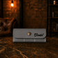 Gray leather wallet with a gold button labeled 'Daniel' on a dark surface with a blurred background.