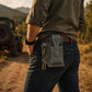Person wearing a gray utility belt with a pouch and keys, standing in a natural outdoor setting with a jeep in the background.