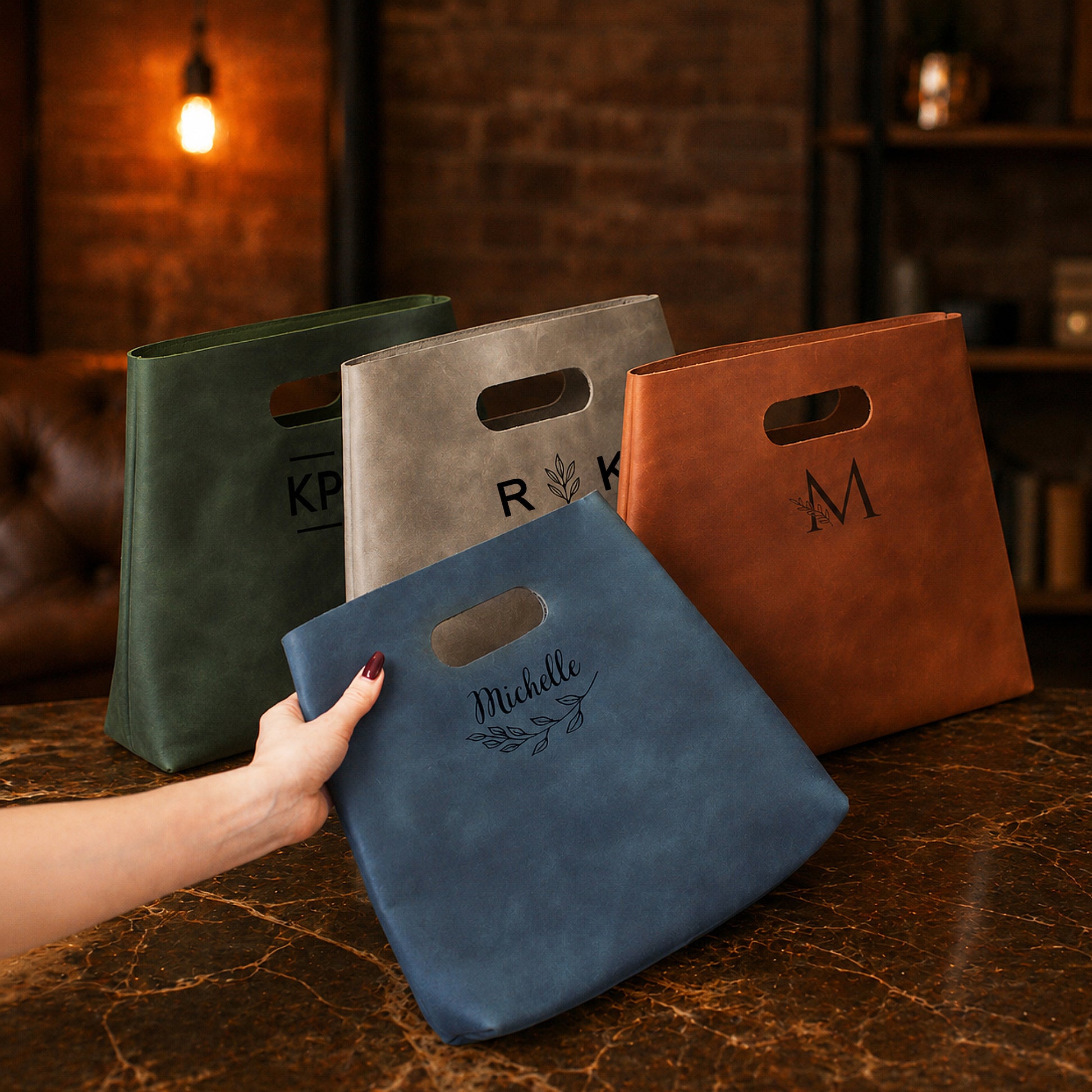 Four leather bags in green, gray, brown, and blue with personalized names on a wooden surface.