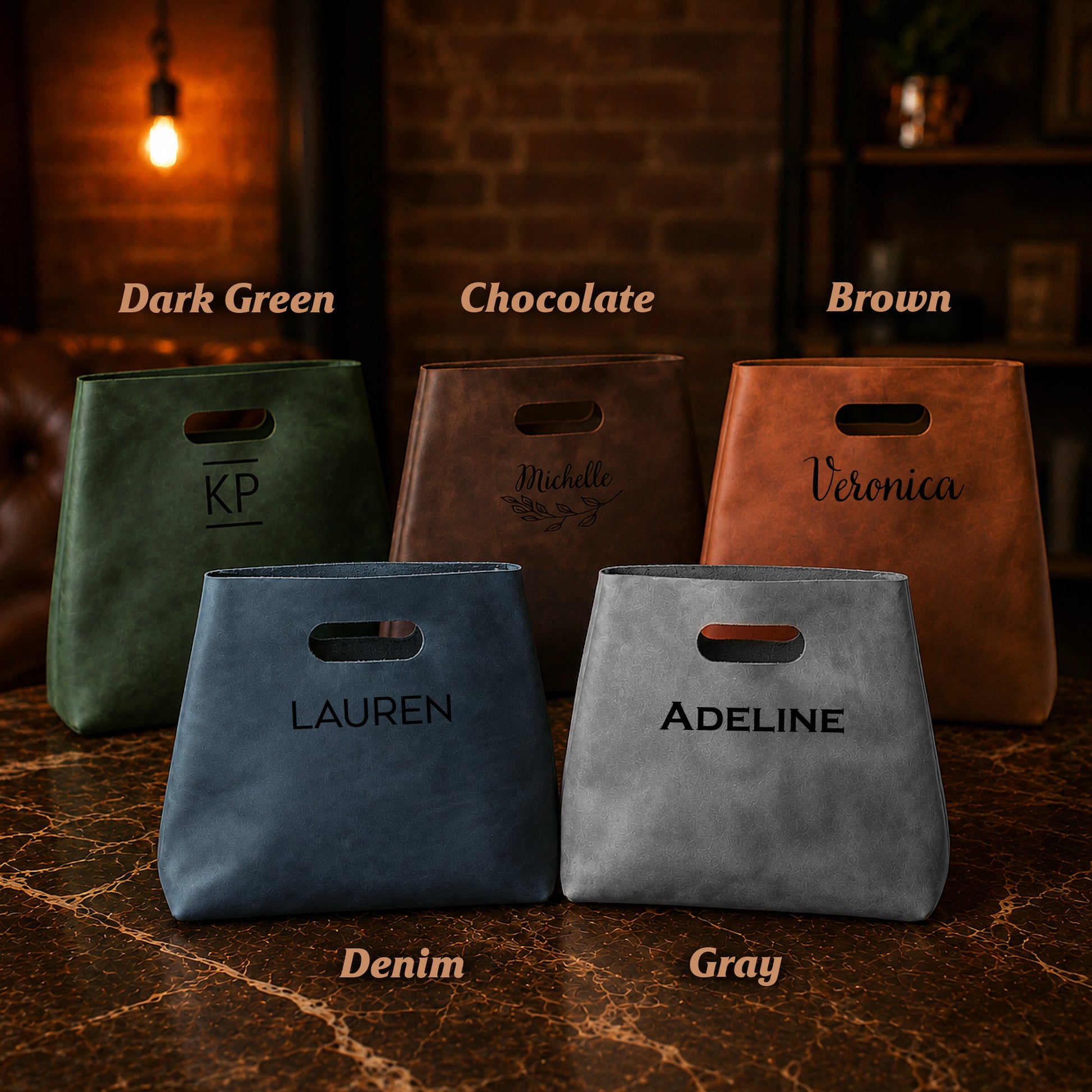 Five leather bags in different colors with names engraved on them, set against a rustic brick wall background.