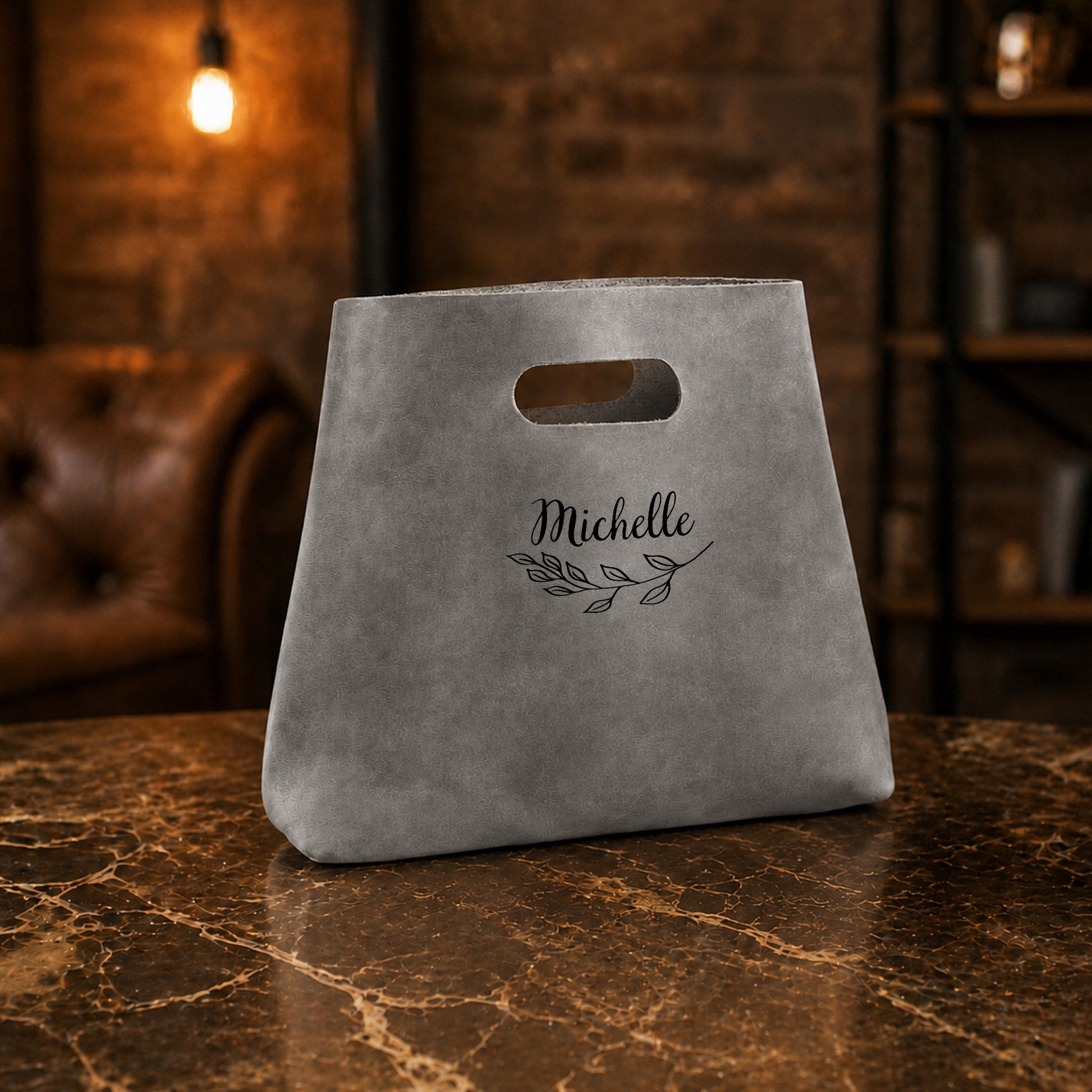 Gray bag with 'Michelle' and a leaf design on a marble surface with a warm-toned background.