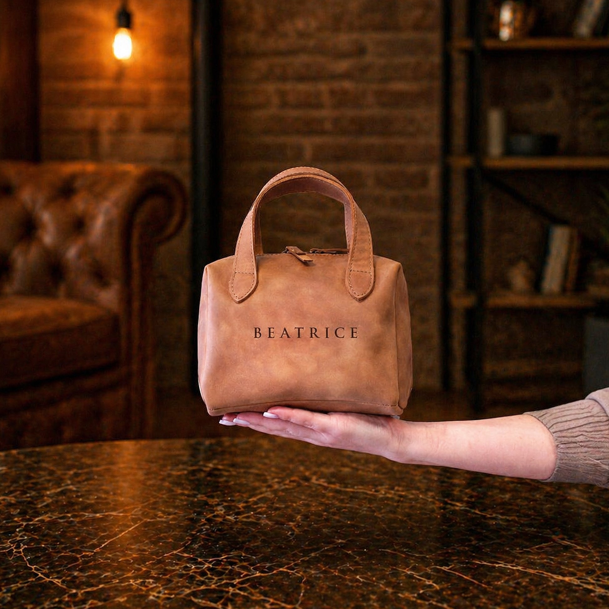 Brown leather handbag with 'BEATRICE' branding held by a person in a warm-toned room.
