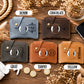 Six small leather card holder wallets arranged on a wooden surface, each labeled with a color name: DENIM, CHOCOLATE, GRAY, TAWNY, and YELLOW. The wallets are shown in blue-gray, dark brown, charcoal, tan, camel, and mustard tones, with monogram and name embossing visible on the front.
