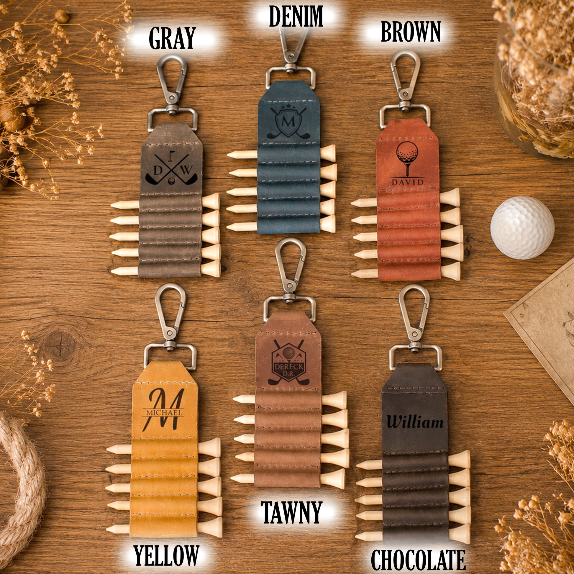 Six personalized golf keychains arranged in a 2x3 grid on a wooden background, each labeled with a color name: Gray, Denim, Brown, Yellow, Tawny, and Chocolate. The keychains feature a leather roll-top strap with multiple pointed wooden spikes, a metal clasp, and engraved names or logos.