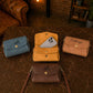 Four suede handbags in blue, orange, and brown on a textured surface with a warm-toned background.